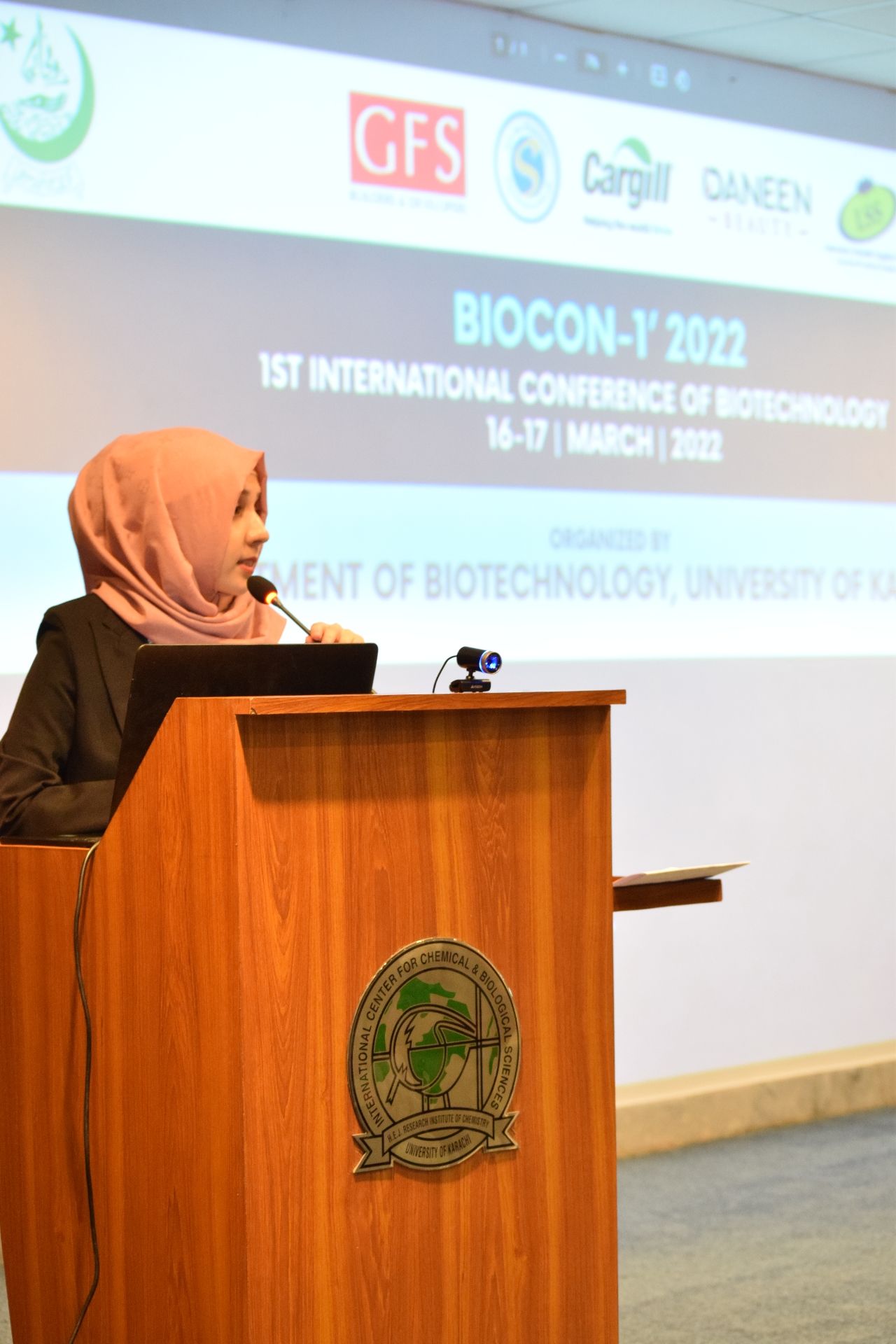 Maha speaking at a conference podium in front of a projected event screen
